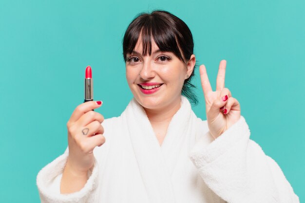 Jonge mooie vrouw die een badjas draagt die een succesvolle overwinning viert en een lippenstift vasthoudt