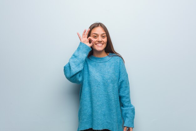 Jonge mooie vrouw die een blauwe sweater vrolijk en zeker draagt die ok gebaar doet