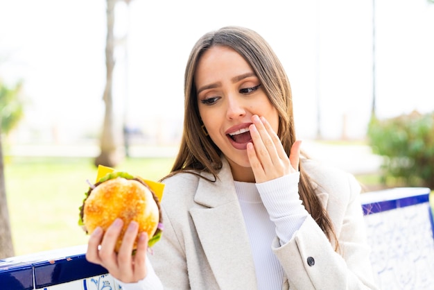 Jonge mooie vrouw die een hamburger buiten vasthoudt met verbazing en geschokte gezichtsuitdrukking