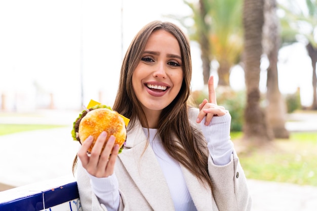 Jonge mooie vrouw die een hamburger in openlucht houdt die op een geweldig idee wijst