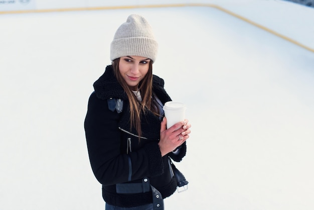 Jonge mooie vrouw die een kopje koffie geeft en op de ijsbaan staat