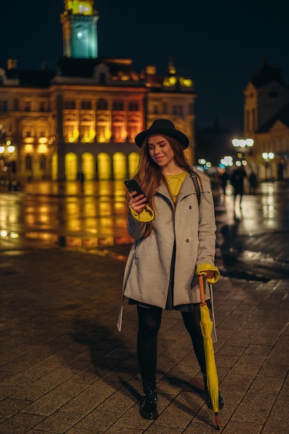 Jonge, mooie vrouw die een smartphone gebruikt en een gele paraplu vasthoudt op een regenachtige nacht