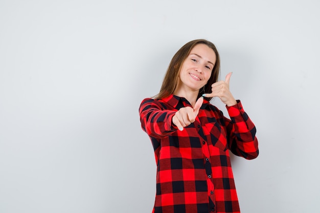 Jonge mooie vrouw die een telefoongebaar maakt, naar de rechterbovenhoek wijst in een casual shirt en er zalig uitziet. vooraanzicht.