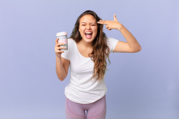 Jonge mooie vrouw die er ongelukkig en gestrest uitziet, zelfmoordgebaar maakt een pistoolteken en houdt een kopje koffie vast