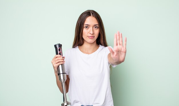 jonge mooie vrouw die er serieus uitziet en open palm toont die een stopgebaar maakt. handmixer concept