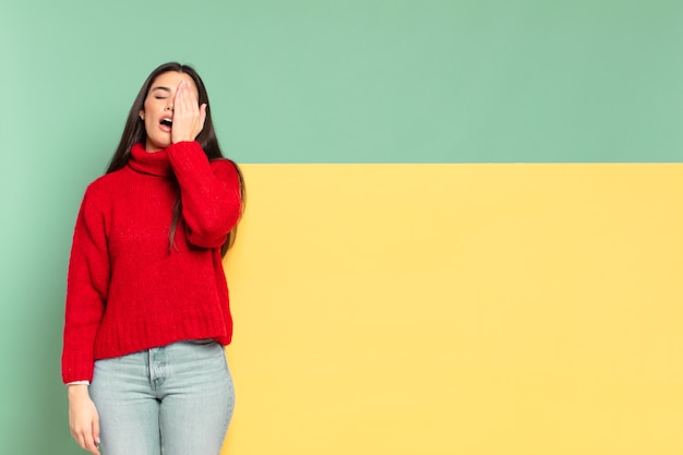 Jonge mooie vrouw die er slaperig, verveeld en geeuwend uitziet, met hoofdpijn en één hand die het halve gezicht bedekt. kopieer ruimte