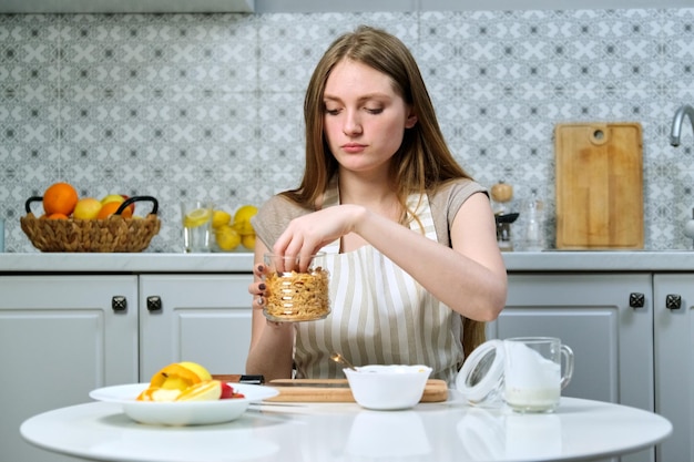 Jonge mooie vrouw die fruitsalade in keuken met appel, sinaasappel, yoghurt, cornflakes en zaden maakt. Gezond eten thuis gekookt, schoonheid en gezondheid van jongeren