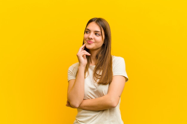 Jonge mooie vrouw die gelukkig en dagdromen of twijfelen glimlachen, die aan de kant tegen oranje muur kijken