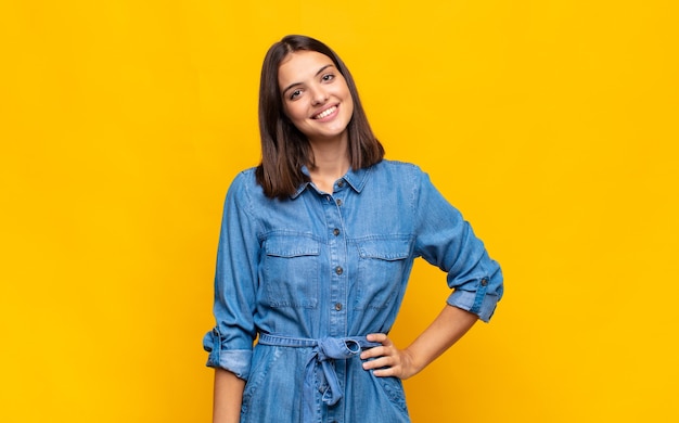 Jonge mooie vrouw die gelukkig glimlacht met een hand op heup en zelfverzekerde, positieve, trotse en vriendelijke houding