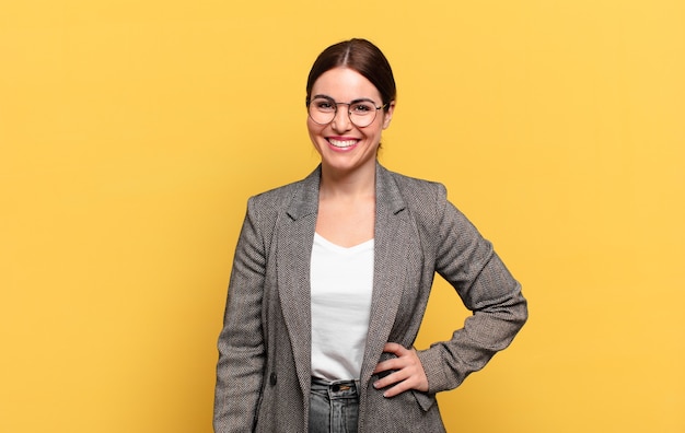 Jonge mooie vrouw die gelukkig glimlacht met een hand op heup en zelfverzekerde, positieve, trotse en vriendelijke houding