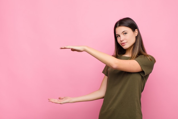 Jonge mooie vrouw die, gelukkig, positief en tevreden glimlachen voelen, of voorwerp of concept op exemplaarruimte tegen roze muur houden tonen of tonen