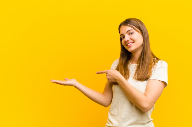 Jonge mooie vrouw die, gelukkig, zorgeloos en tevreden glimlacht voelt, wijzend op concept of idee op exemplaarruimte aan de kant over oranje muur