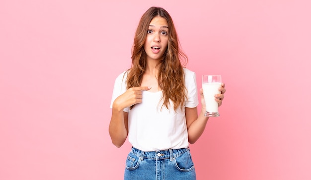 Jonge mooie vrouw die geschokt en verrast kijkt met wijd open mond, wijzend naar zichzelf en een glas melk vasthoudend