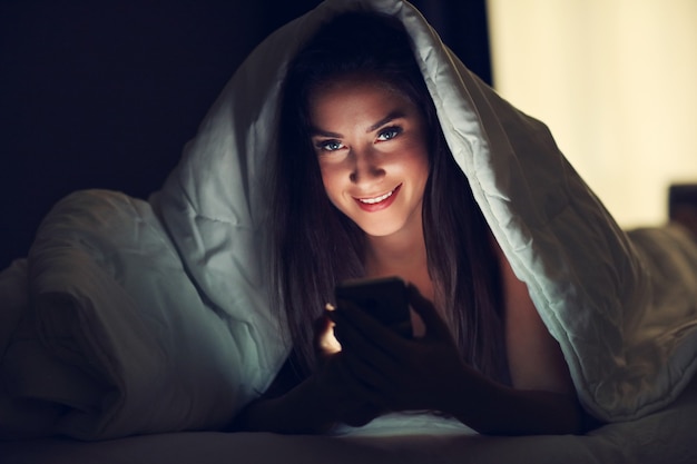 Jonge mooie vrouw die in bed met smartphone rust