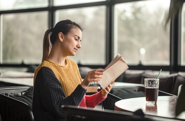 Jonge mooie vrouw die in oranje sweater interessant boek in koffie lezen