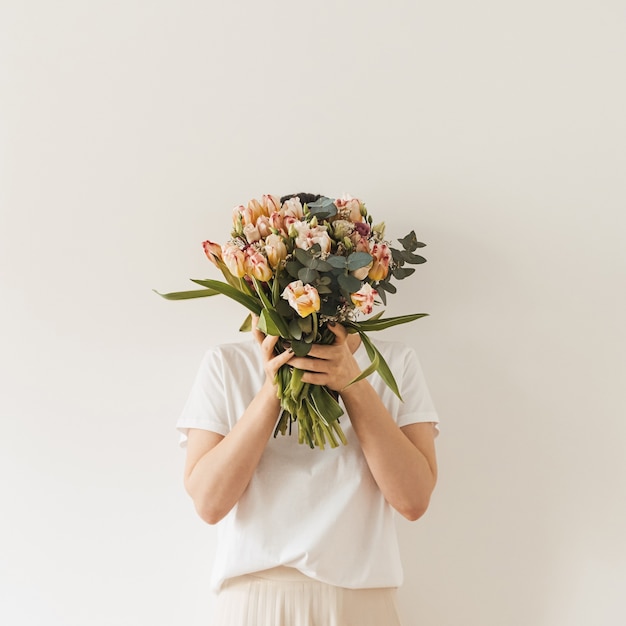 Jonge mooie vrouw die in witte blouse het boeket van tulpbloemen in handen houdt tegen witte muur