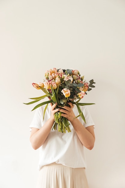 Jonge mooie vrouw die in witte blouse het boeket van tulpbloemen in handen houdt tegen witte muur