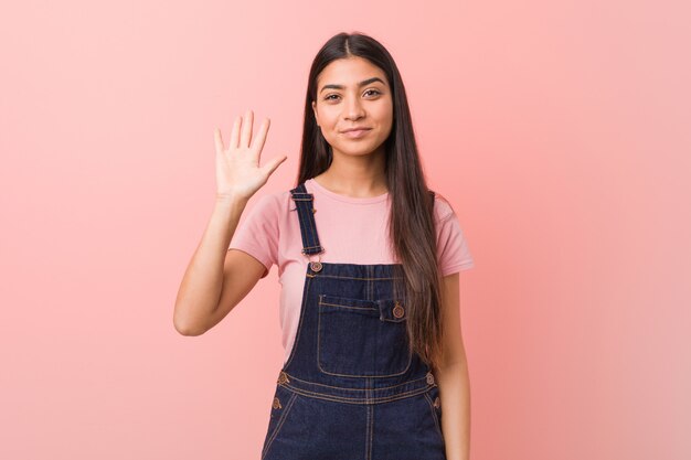 Jonge mooie vrouw die jeansbroekmassa draagt die vrolijk tonend nummer vijf met vingers glimlacht