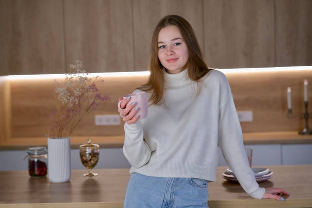 Jonge mooie vrouw die koffie of thee drinkt uit de beker in de keuken in een trui die lacht