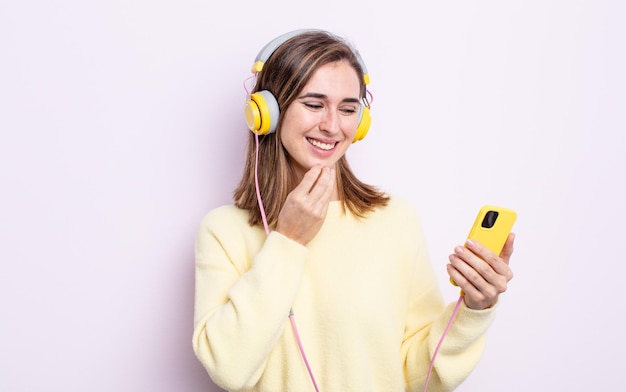 Jonge mooie vrouw die lacht met een gelukkige, zelfverzekerde uitdrukking met de hand op de kin. koptelefoon en telefoon concept