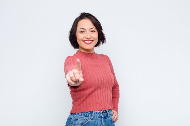 Jonge mooie vrouw die lacht trots en vol vertrouwen het maken van nummer één pose triomfantelijk, het gevoel als een leider tegen de witte muur