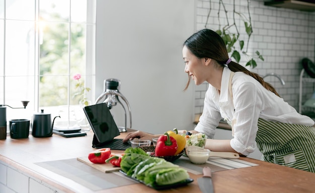 Jonge mooie vrouw die laptopcomputer gebruikt die zoekt en leert voor het koken van gezond voedsel van verse groenten en fruit in de keukenruimte