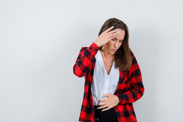 Jonge mooie vrouw die lijdt aan buikpijn, met vinger op voorhoofd in casual outfit en er pijnlijk uitziet. vooraanzicht.