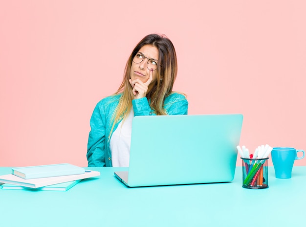 Jonge mooie vrouw die met een laptop met een geconcentreerde blik werkt, zich afvragend met een twijfelachtige uitdrukking, omhoog kijkend en opzij