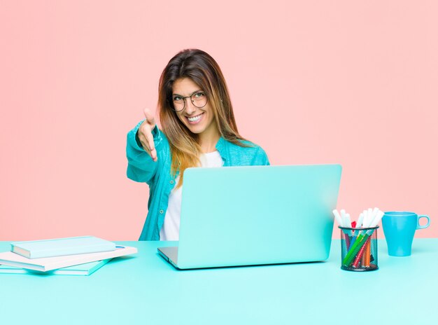 Jonge mooie vrouw die met laptop glimlachen werkt, die gelukkig, zeker en vriendschappelijk kijkt, die een handdruk aanbiedt om een overeenkomst te sluiten