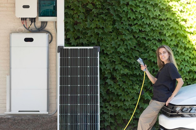 Jonge, mooie vrouw die naar de camera kijkt en glimlacht op een witte elektrische auto met een stekker en een zonne-energie