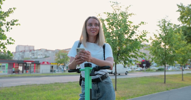 Jonge mooie vrouw die naast een elektrische scooter staat terwijl ze haar smartphone gebruikt Moderne architectuur op de achtergrond