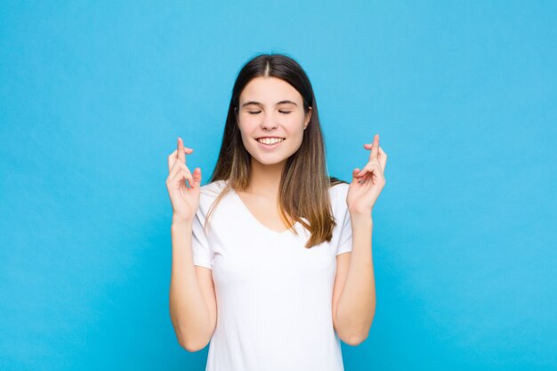 Jonge mooie vrouw die nerveus en hoopvol is, vingers kruist, bidt en hoopt op geluk tegen blauwe muur