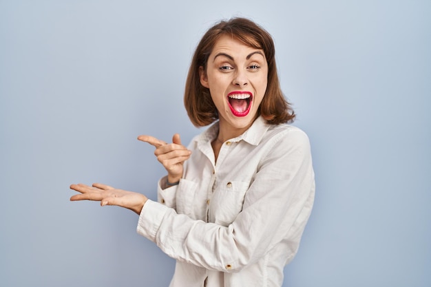 Jonge mooie vrouw die nonchalant over blauwe achtergrond staat verbaasd en glimlachend naar de camera terwijl ze met de hand presenteert en met de vinger wijst.