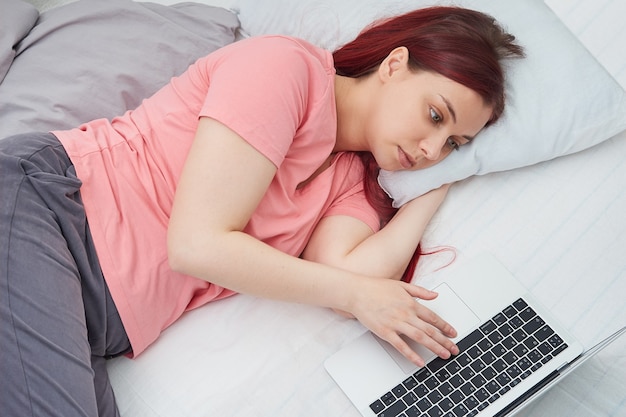 Jonge mooie vrouw die op afstand aan laptop werkt terwijl ze in bed ligt.