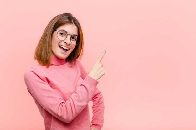 Jonge mooie vrouw die opgewekt en verrast het richten aan de kant en naar omhoog kijken om ruimte tegen roze muur te kopiëren