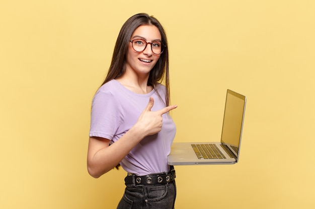 Jonge mooie vrouw die opgewonden en verrast kijkt en naar de zijkant en naar boven wijst om ruimte te kopiëren. laptop concept