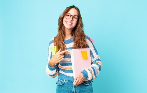 Jonge mooie vrouw die opgewonden en verrast kijkt en naar de zijkant wijst met een tas en boeken vasthoudt