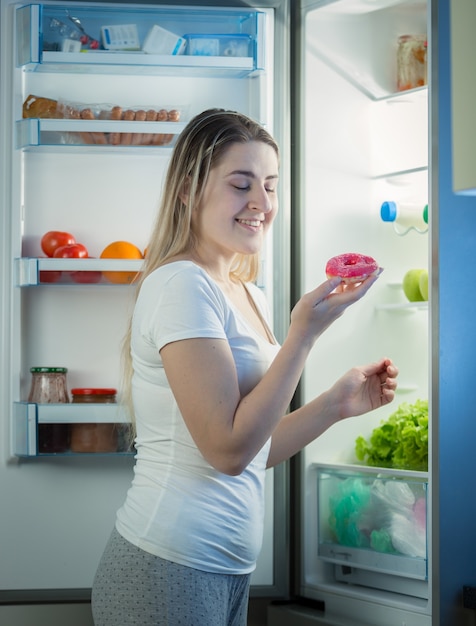 Jonge mooie vrouw die 's avonds laat donut uit de koelkast haalt