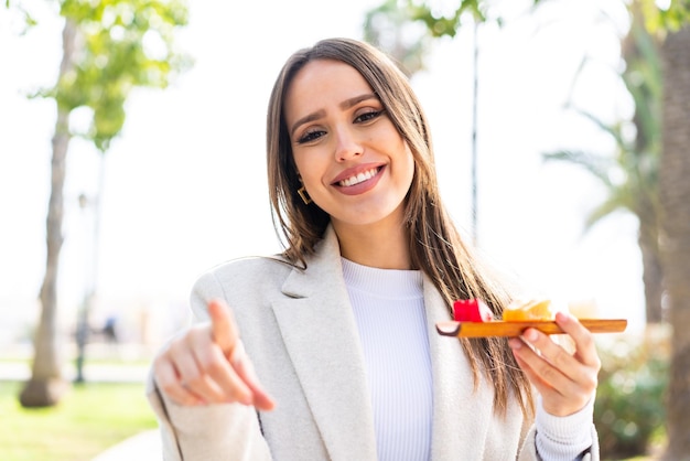 Jonge mooie vrouw die sashimi buiten vasthoudt, wijst met een zelfverzekerde uitdrukking naar je