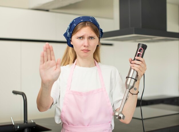 Foto jonge mooie vrouw die serieus kijkt met open handpalm en stopgebaar maakt, chef-kok en handblenderconcept