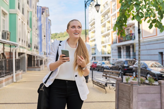 Jonge mooie vrouw die smartphone gebruikt die langs de stadsstraat loopt, zakenvrouw in een wit jasje met laptoptas