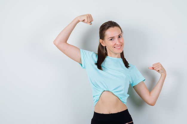 Jonge mooie vrouw die spieren van armen in t-shirt toont en er trots uitziet, vooraanzicht.