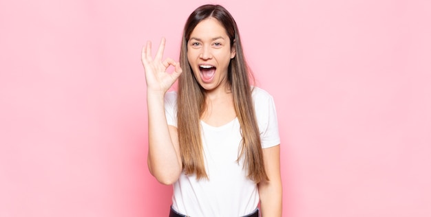 Jonge mooie vrouw die succesvol en tevreden voelt, glimlachend met wijd open mond, goed teken met hand maakt