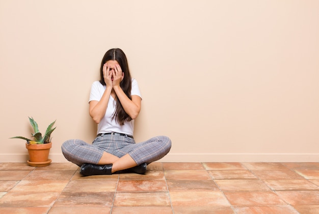 Jonge mooie vrouw die verdrietig, gefrustreerd, zenuwachtig en depressief voelt, gezicht bedekkend met beide handen, huilend zittend op een vloer