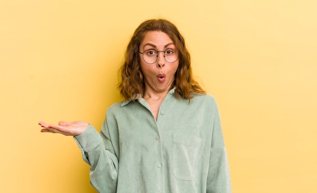 Jonge mooie vrouw die verrast en geschokt kijkt met open mond terwijl ze een object vasthoudt met een open hand aan de zijkant