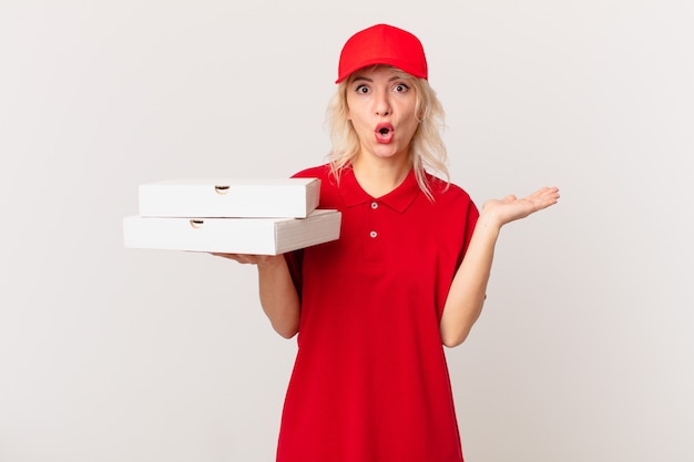 Jonge mooie vrouw die verrast en geschokt kijkt, met open mond terwijl ze een object vasthoudt. pizza bezorgen