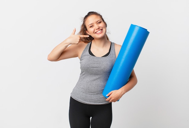 Jonge mooie vrouw die vol vertrouwen glimlacht, wijst naar haar eigen brede glimlach en een yogamat vasthoudt