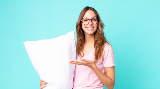 Jonge mooie vrouw die vrolijk lacht, zich gelukkig voelt en een concept toont dat een pyjama draagt en een kussen vasthoudt