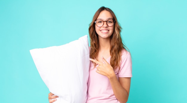 Jonge mooie vrouw die vrolijk lacht, zich gelukkig voelt en naar de zijkant wijst met een pyjama aan en een kussen vasthoudt