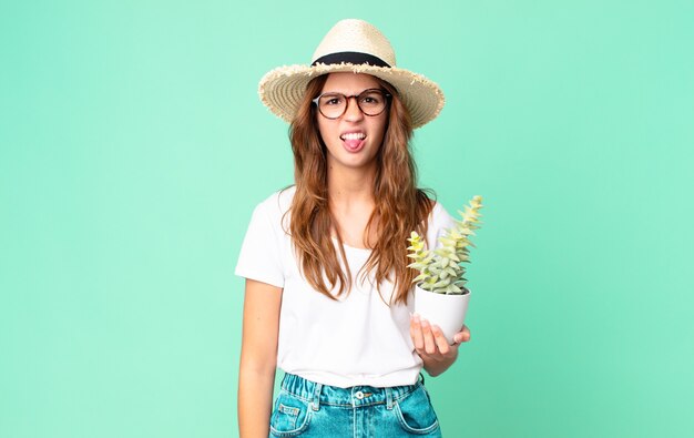 Jonge mooie vrouw die walgt en geïrriteerd voelt en tong uitsteekt met een strohoed en een cactus vasthoudt